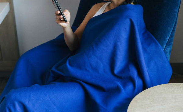 A Large Shepherd's Wool Utility Cloth in Neela Blue being worn as a blanket while a person rests in a blue velvet chair reading something on their phone.